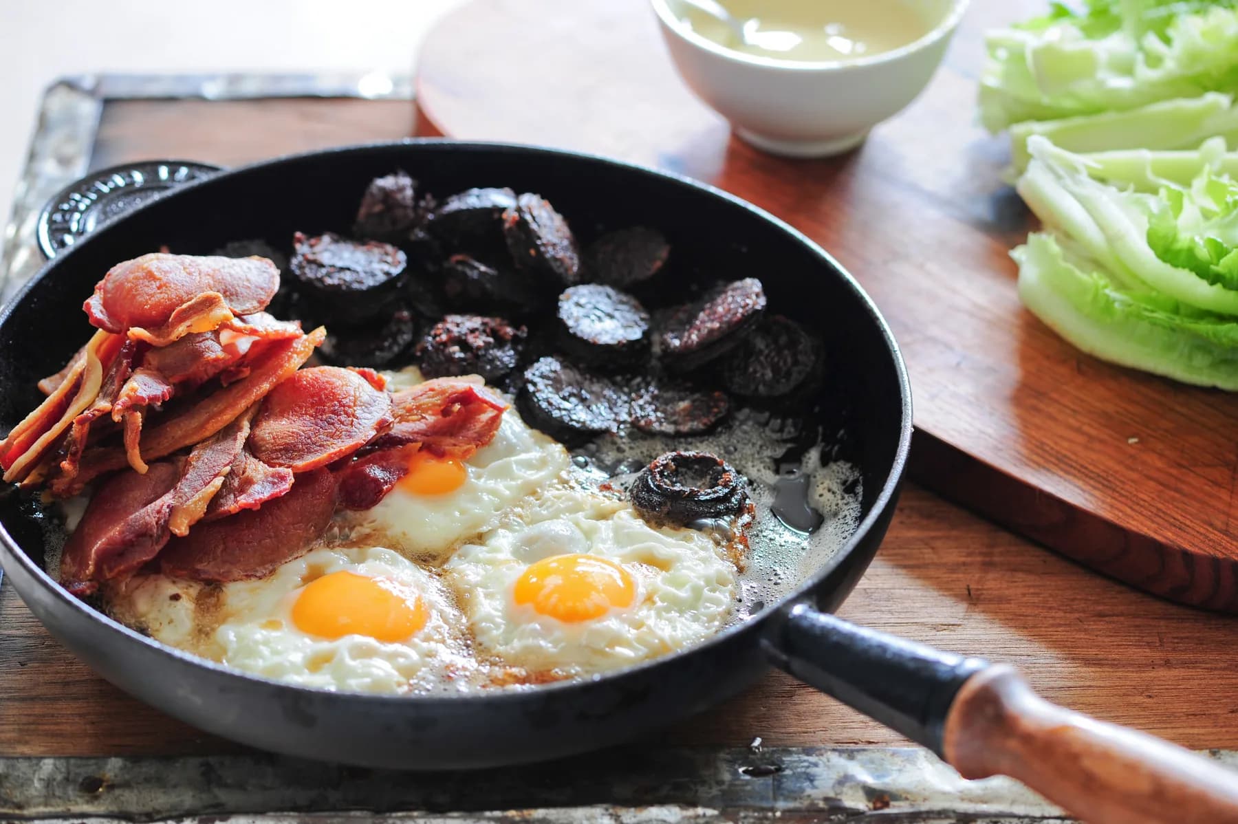 A pan of a full Irish breakfast cooking: back-bacon rashers, sliced black pudding, and fried eggs