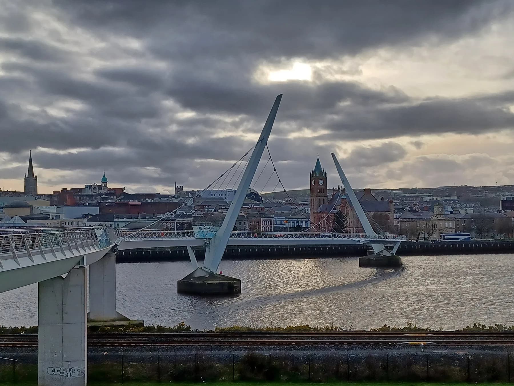 The Peace Bridge curving across the River Foyle in Derry, with the city skyline and Guildhall clock tower behind under a grey sky