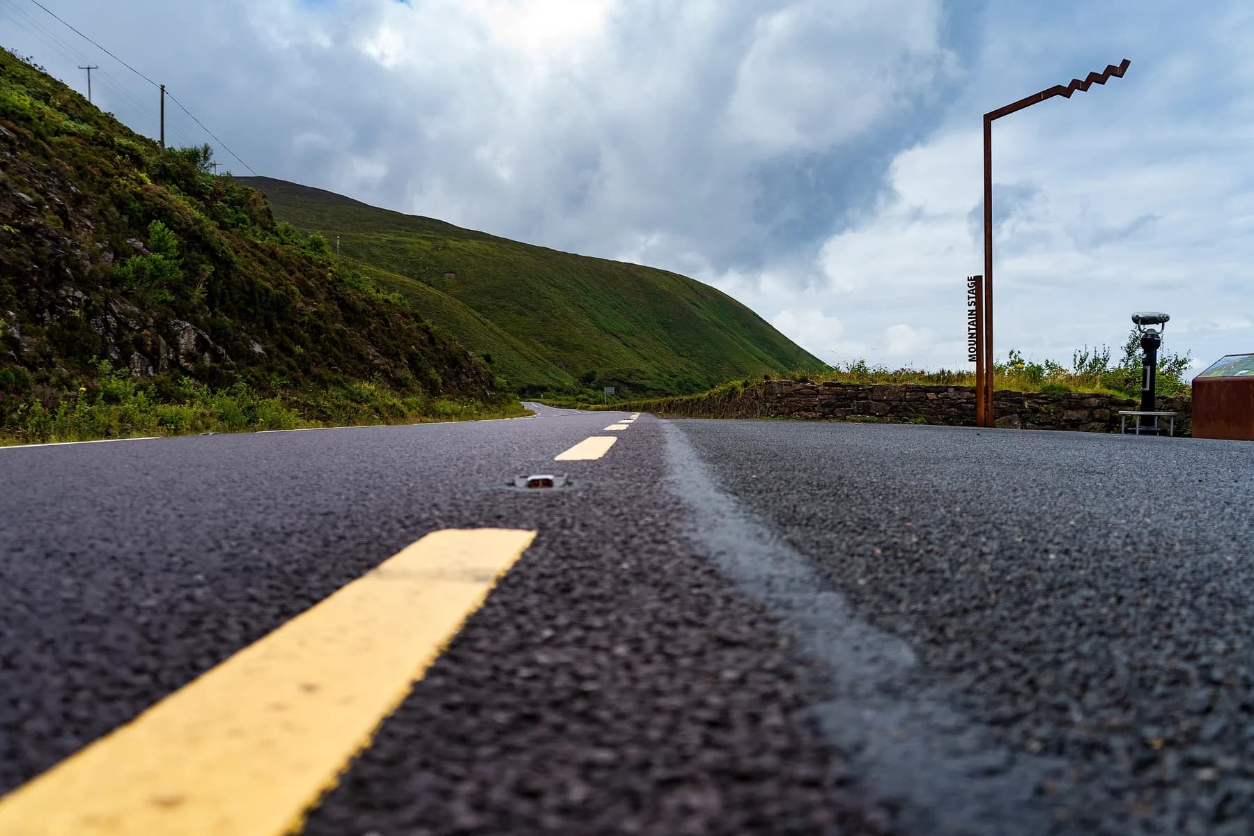 A Wild Atlantic Way coastal road with the route's zigzag marker at a Discovery Point, green mountains and a moody sky behind